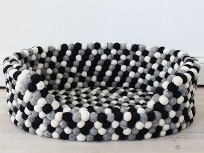 A round grey felt pet bed made of small wool balls arranged in a circular pattern, sitting on light wooden flooring against a white wall